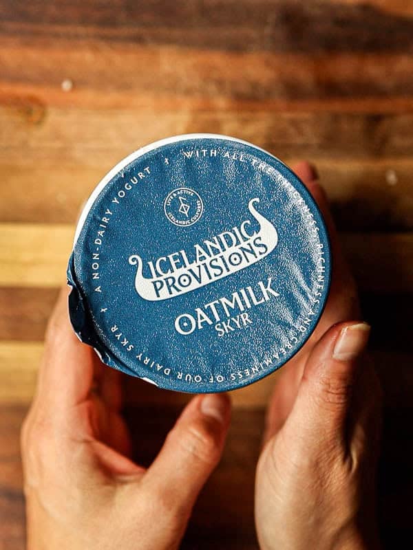 Close-up of hands holding a container with a blue lid labeled Icelandic Provisions Oatmilk Skyr. The lid is slightly pulled back on one side. The background is a wooden surface.